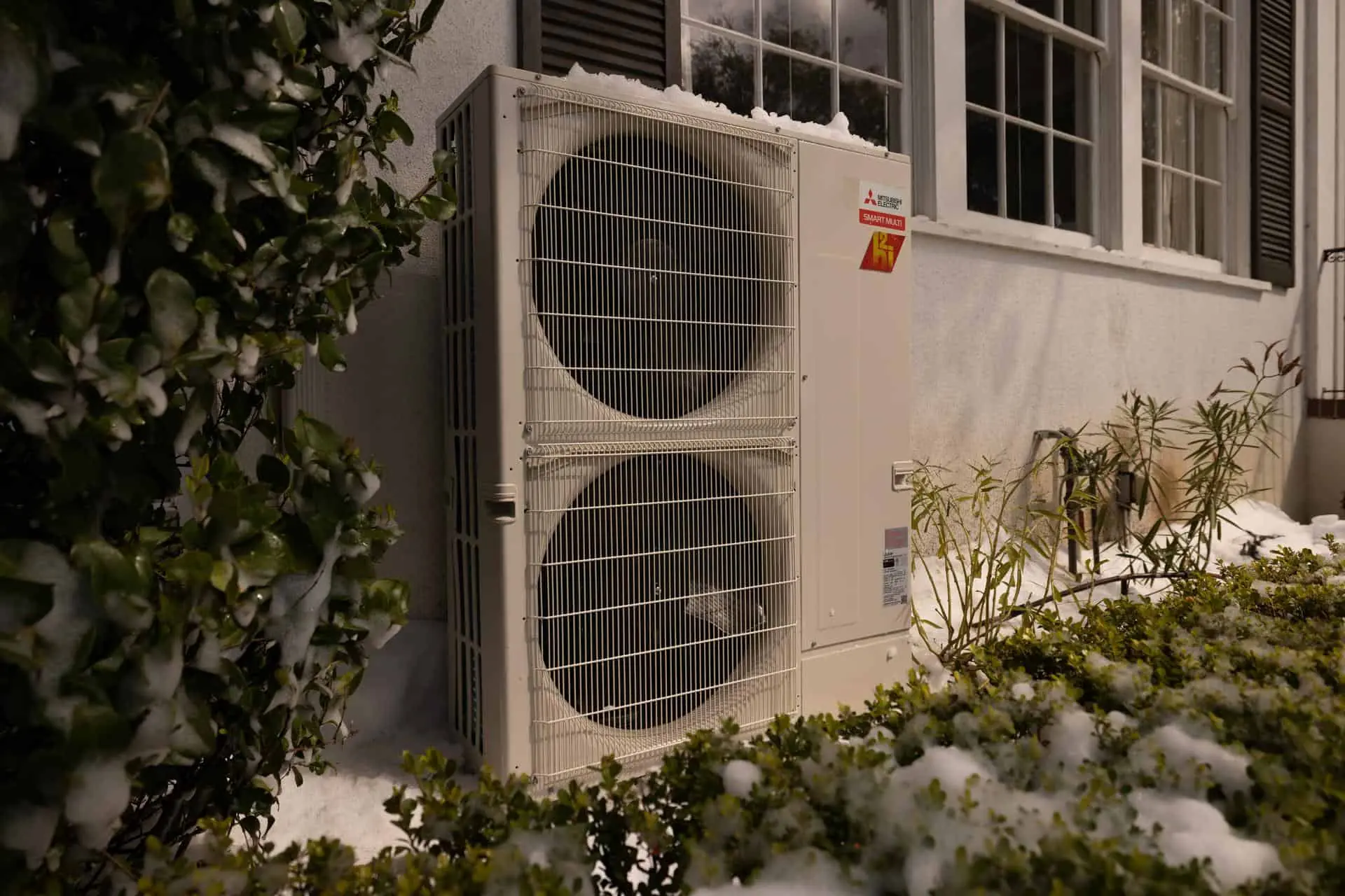 A Mitsubishi heat pump outside a Denver home, the shrubs around it covered in snow.