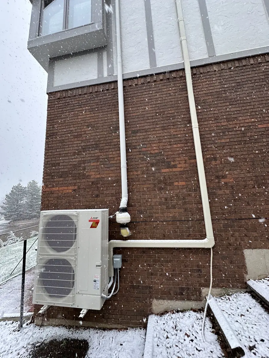 A Mitsubishi cold-climate heat pump running in falling snow against a brick wall.